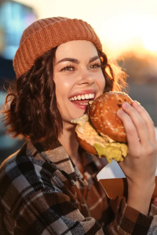 Woman eating burger outdoors with beanie and plaid jacket, bright smile in golden hour glow, candid lifestyle portrait conveying authenticity, mindful living and emotional storytelling.