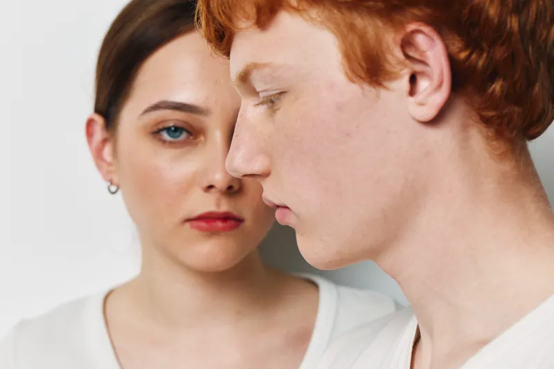 Young man and woman portrait with serious expressions closeup, studio background and casual clothing, focusing on natural skin texture and facial details in soft lighting.