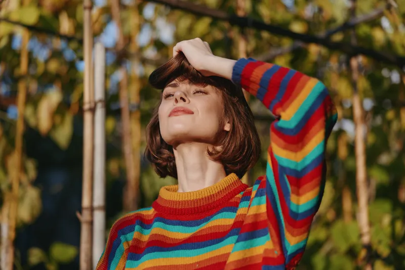 Woman portrait in rainbow sweater basking in warm sunlight outdoors, smiling with eyes closed and hand shading forehead, cozy knit fashion and relaxed autumn garden mood.