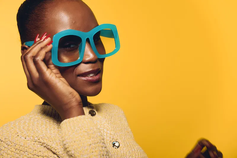 Confident black woman with dark skin wearing oversized blue glasses and a beige knitted sweater poses against a yellow background. Concept of fashion, style, and modern accessories.
