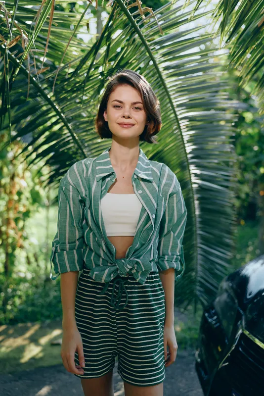 Casual young woman stands outdoors among tropical palms, wearing a green striped shirt tied at the waist and matching striped shorts with a light crop top.