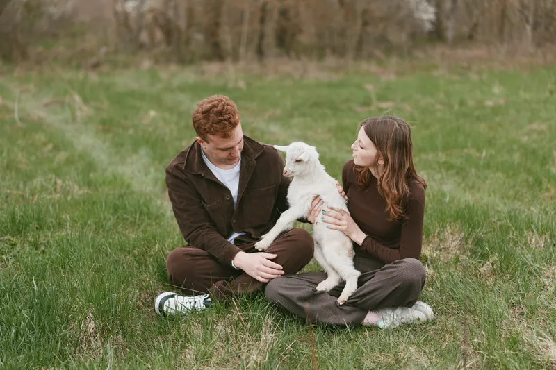 Happy couple shares a moment with their small white dog in a grassy park, sitting close and smiling, enjoying a leisurely outdoor afternoon together, with relaxed casual style and friendly mood.