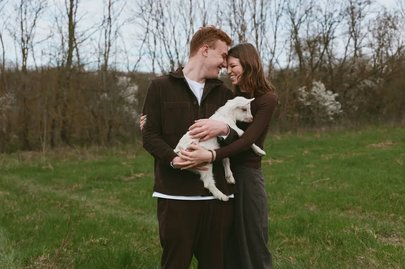 A young couple embraces tenderly in a grassy field while holding a small white baby goat, sharing a joyful moment during a casual outdoor lifestyle shoot in early spring.