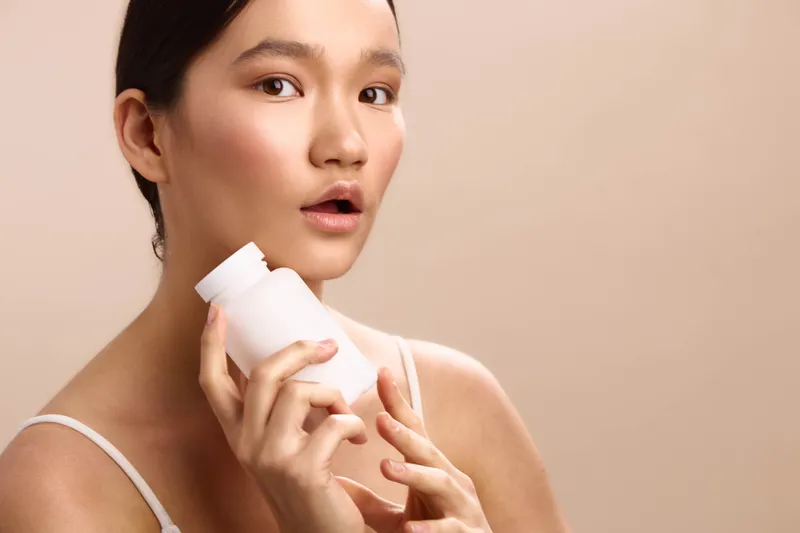 asian woman with a skincare bottle in a soft studio setting, presenting a calm, healthy beauty moment as she gazes slightly off camera with a gentle, confident expression.