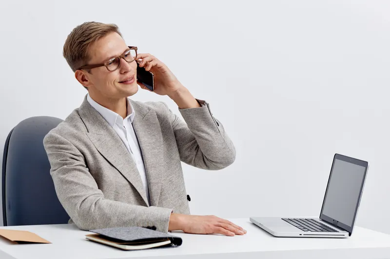 Smiling young caucasian man talking smartphone sitting workspace with laptop notebook modern business casual fashion digital nomad remote work concept