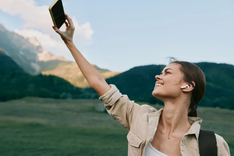 Smiling woman taking a selfie outdoors with mountains and green fields in the background. A lifestyle moment emphasizing exploration, wellbeing, and cheerful energy.