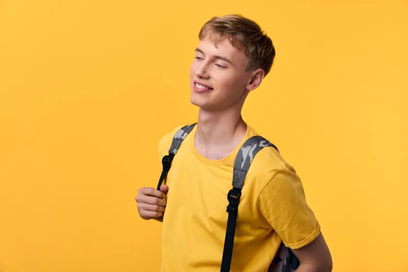 Youthful student with backpack smiles against a bright yellow background, casual attire suitable for travel and daily life, projecting confidence, energy, and a positive mood for learning and new