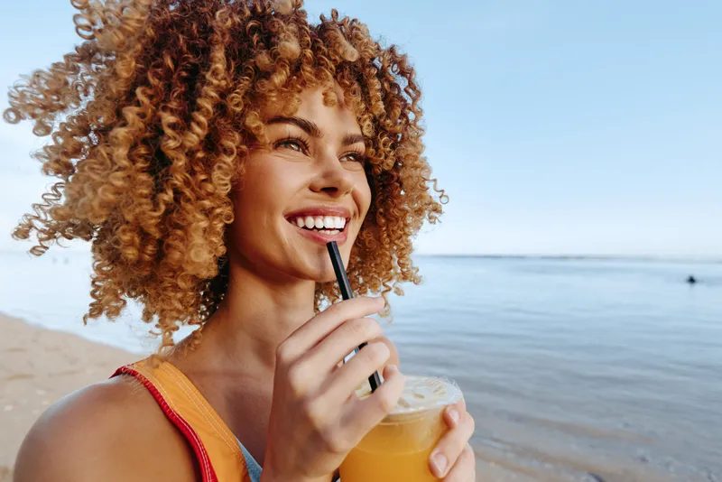 Young woman with curly hair and a bright smile enjoys a refreshing drink by the calm sea on a sunny day, wearing a red swimsuit and feeling relaxed and happy.