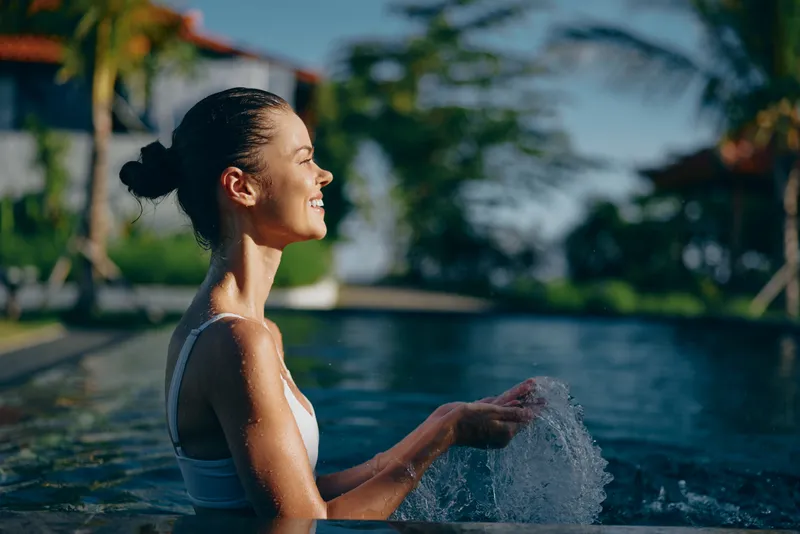 Happy woman in a sunlit pool enjoys refreshing water moment, smiling and relaxed, embodying outdoor leisure and summer lifestyle with calm, bright surroundings and tropical breeze, creating a candid,