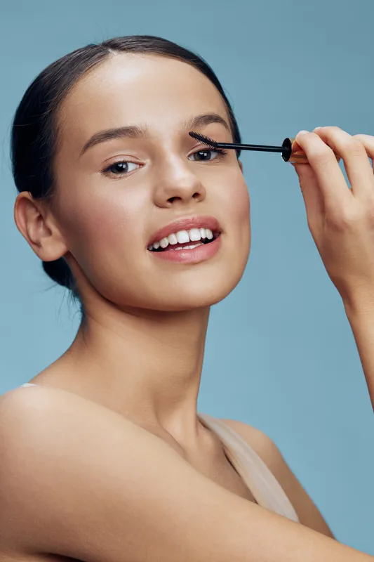 Young woman applying mascara on her eyelashes, showcasing beauty routine with a bright blue background, exuding confidence and positivity in a minimalistic style