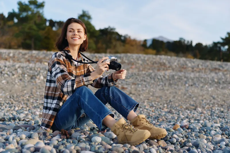 Woman with camera in outdoor casual photography, sitting on pebbles in plaid shirt and boots, smiling under natural light by a rocky shore, capturing a candid moment during a tranquil nature shoot.