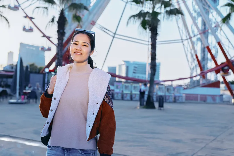 Young woman posing outdoors in a casual spring outfit, light sweater and jacket, palm trees and modern architecture behind, bright daylight street style for travel, fashion, and lifestyle photography