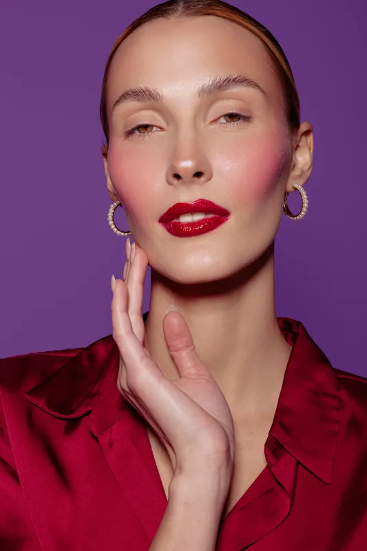 Close-up portrait of a confident woman with glossy red lips and gold hoop earrings wearing a red satin blouse against purple background. Concept of beauty, fashion, and elegance.