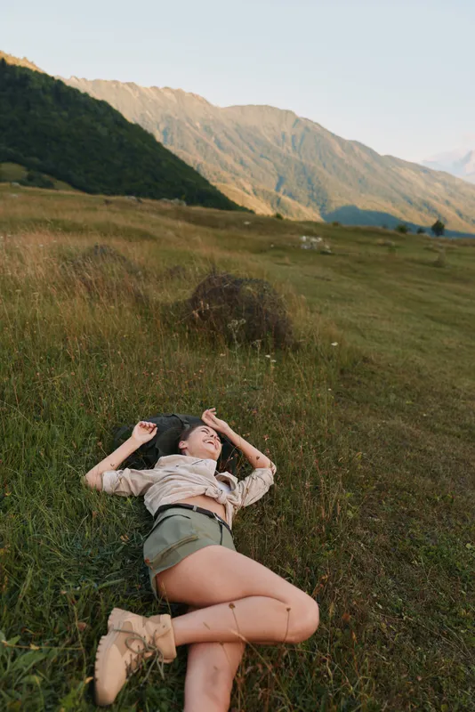 Relaxed woman lying in a field with mountains in the background, casual countryside vibe, sunlit calm, carefree lifestyle moment.