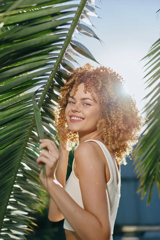 Smiling woman with curly hair enjoying sunny tropical outdoors, surrounded by green palm leaves and natural light, radiating happiness and warmth.
