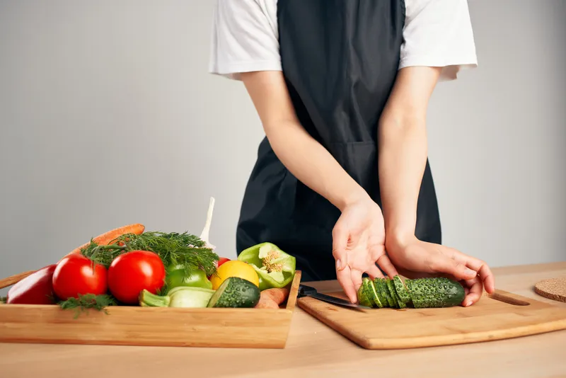 Женщина в черном фартуке нарезает овощи на кухне, домохозяйка готовит еду