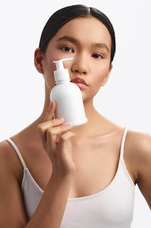 Asian woman holds a white skincare bottle near her face in a clean studio setting, exuding calm confidence and focus on personal care, health, and beauty in a minimal, well lit composition.