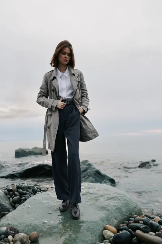 Young woman in a long coat and dark pants stands on rocky shore near ocean under cloudy sky. Fashion portrait with casual style and moody natural light by the sea landscape.