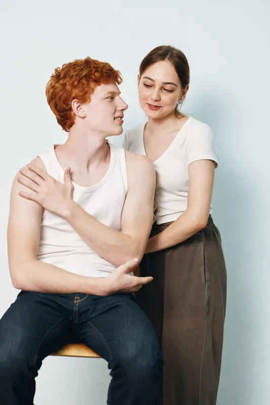 Young couple with man sitting on stool and woman standing behind him, showing affection and closeness in casual clothes on light background.