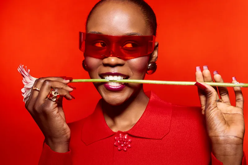 Stylish dark skinned African American woman wearing red sunglasses and red top bites a yellow flower stem, posing against red background. Concept of fashion, beauty, and vibrant color contrast.