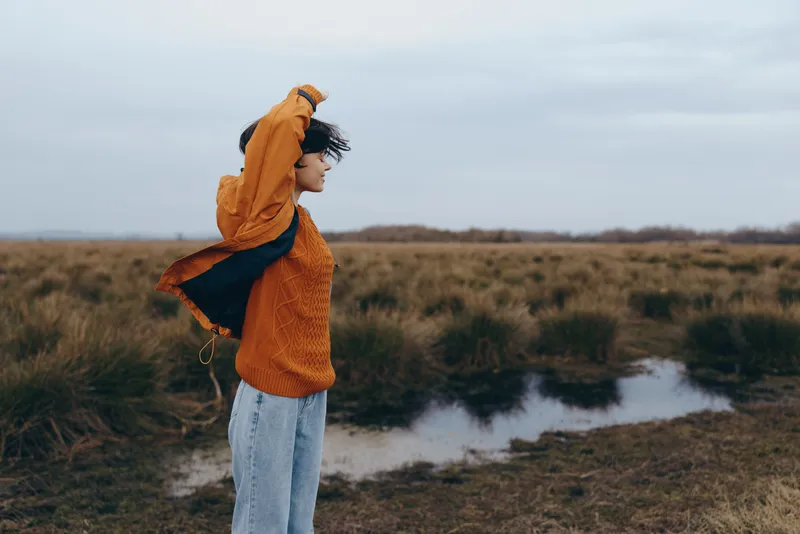 Woman lifestyle outdoors, hooded orange jacket and jeans, standing near wet field with water pool, side profile looking away, calm mood, cloudy sky, natural earthy colors