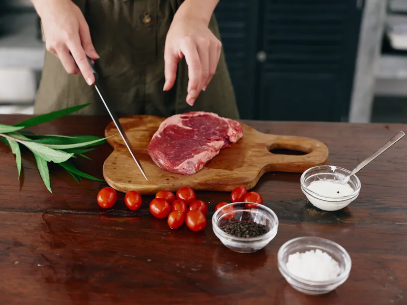 Женщина с ножом в руке нарезает мясо для стейка на жарку на кухне