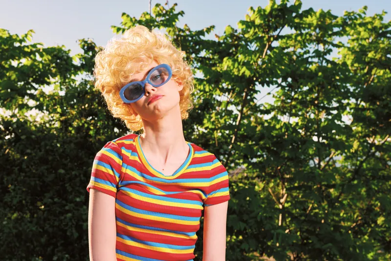 Smiling woman with curly hair wearing a striped colorful shirt and oversized sunglasses. Outdoors in bright daylight with green foliage, retro vibe and playful fashion mood.