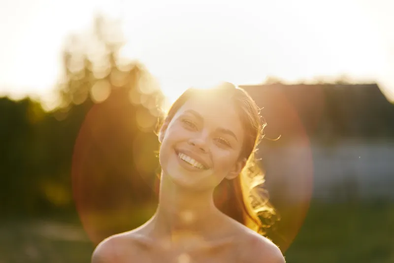 Young woman smiling against a sunlit backdrop in a natural setting, exuding happiness and warmth, showcasing a carefree summer vibe with a glowing effect