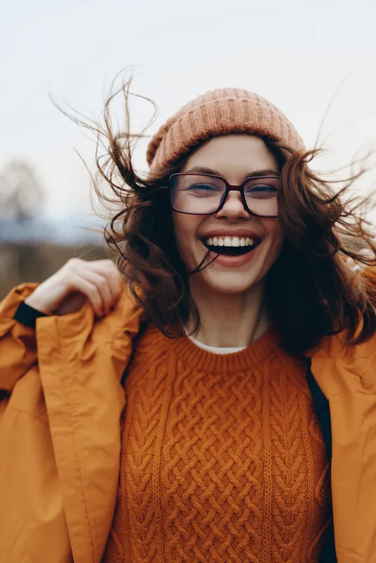 Cheerful woman lifestyle outdoors, laughing in the wind with eyes closed, wearing knit sweater and beanie, round glasses, warm autumn sun, breeze and flowing hair, blurred park
