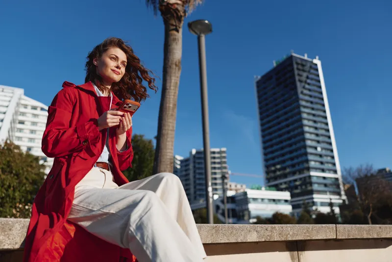 Young woman in a bright red coat sits on a concrete ledge outdoors, smiling while scrolling her smartphone with a modern city skyline and clear blue sky behind.