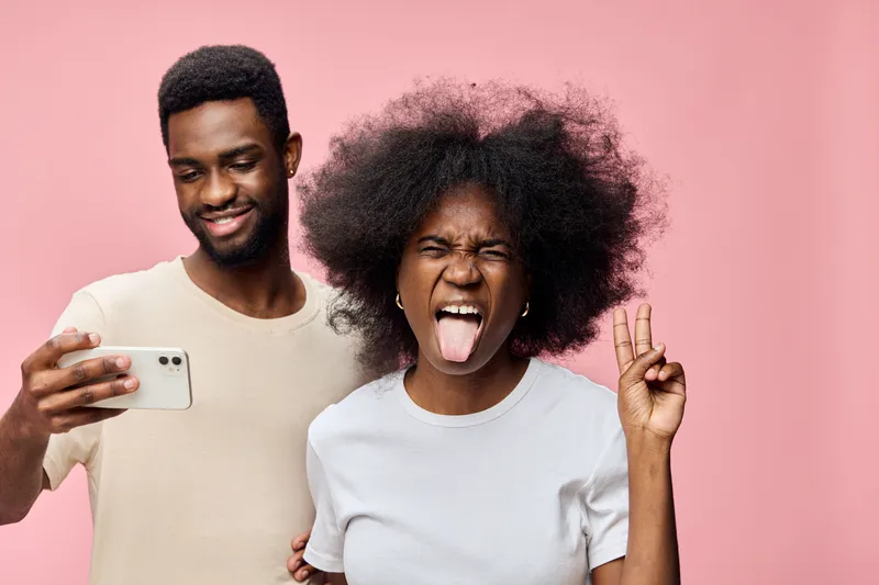 Joyful young couple taking a selfie against a pink background, showcasing vibrant emotions and playful poses, perfect for social media and lifestyle concepts