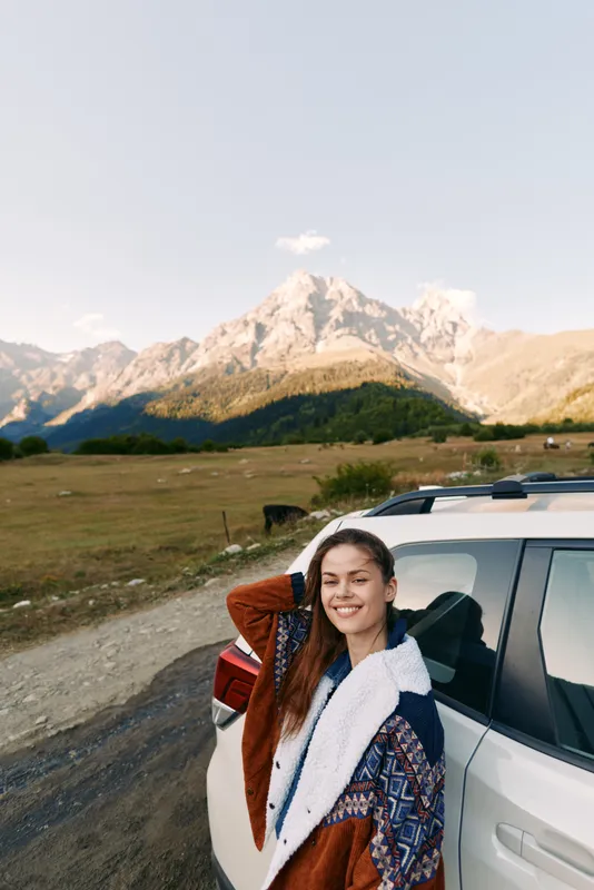 Woman with car on mountain roadtrip, smiling and leaning against vehicle near rugged landscape. Young traveler in cozy jacket enjoying outdoor scenery and clear sky.