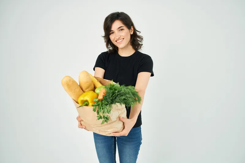 Весёлая женщина с пакетом продуктов, овощами, здоровой пищей на светлом фоне