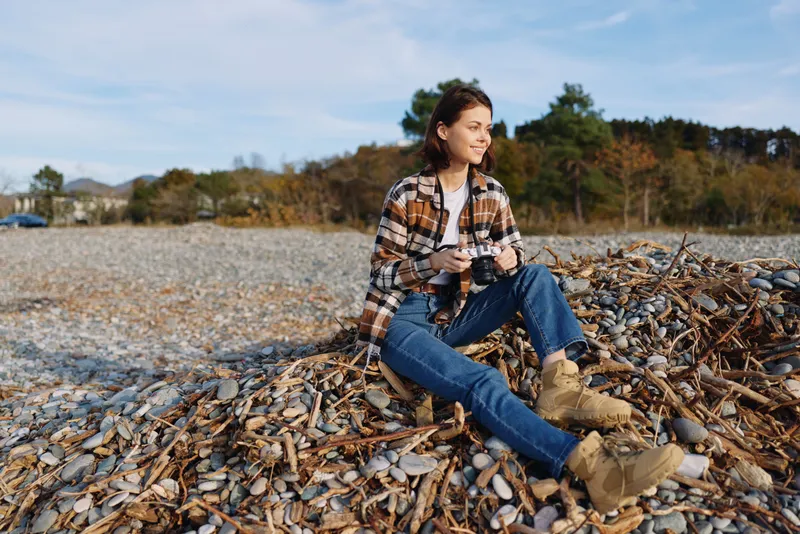 Woman sits on a pile of dried leaves at a rocky beach, dressed in a plaid shirt, jeans and boots, enjoying a calm outdoor moment in autumn light.