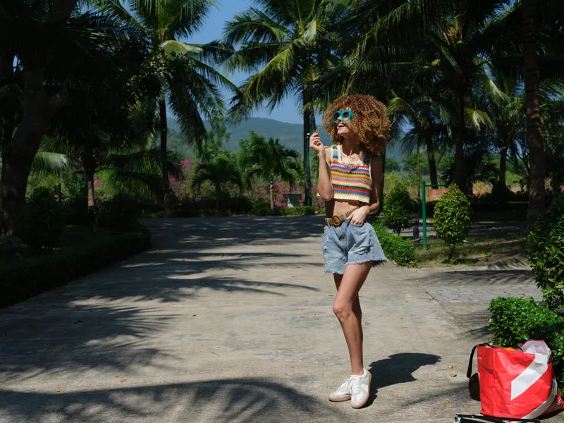 smiling woman in summer clothes enjoying tropical garden, wearing sunglasses and denim shorts, vibrant outdoor setting, sunny day