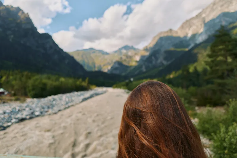 A traveler stands near a flowing river in a dramatic mountain valley, facing away from the camera. The scene conveys solitude, exploration, and a sense of wide open nature.