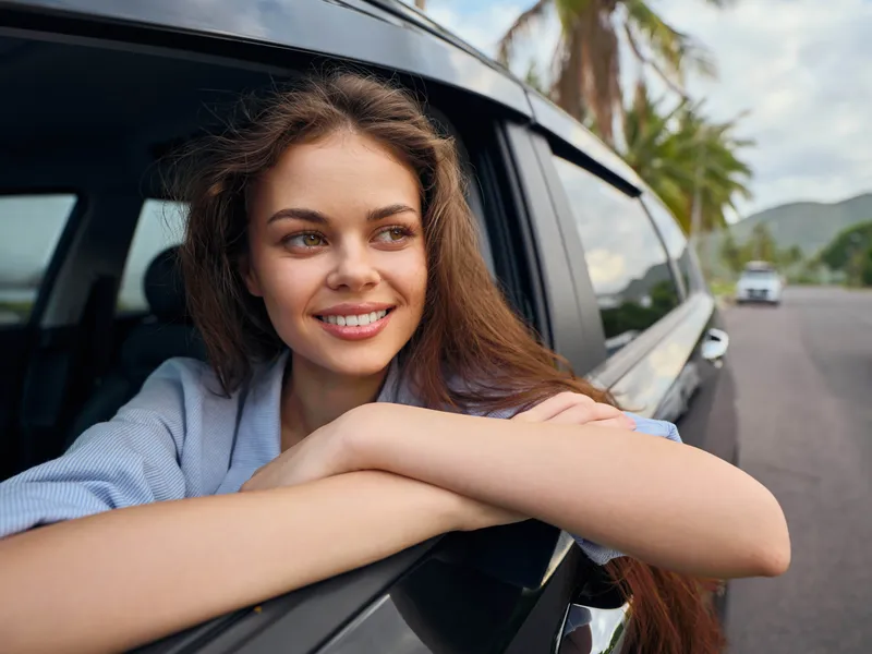 Улыбающаяся молодая женщина сидит на пассажирском сиденье автомобиля на живописной дороге, вдоль которой растут пальмы