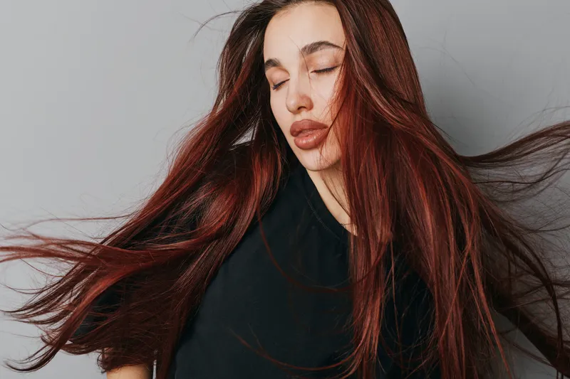 Woman with long red hair and closed eyes wearing black shirt on white background. Portrait of female model with flowing hair and natural makeup in studio for beauty and fashion.