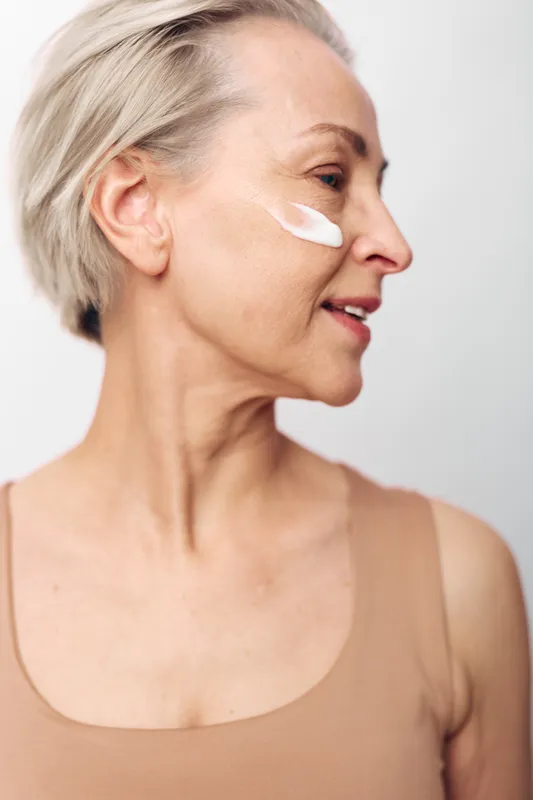 Mature woman with short hair applying face cream on cheek. Close-up profile on light background. Concept of skincare, aging skin care, wellness, and self-care routine.