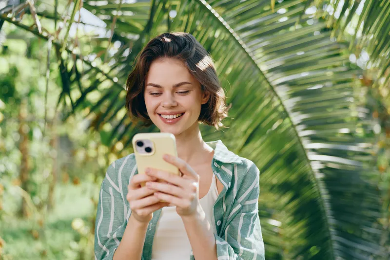Young woman smiling while looking at a smartphone in a sunny outdoor setting with lush green palm leaves.