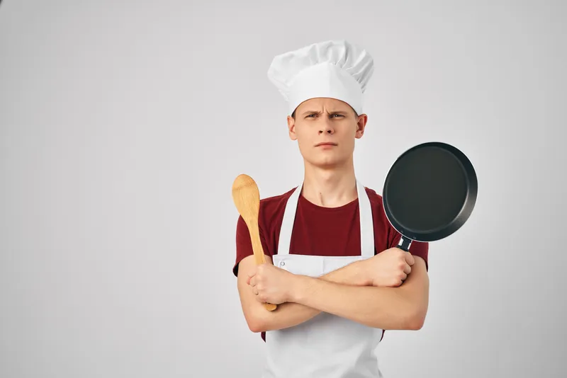 Повар в колпаке держит в руках сковороду, готовя еду