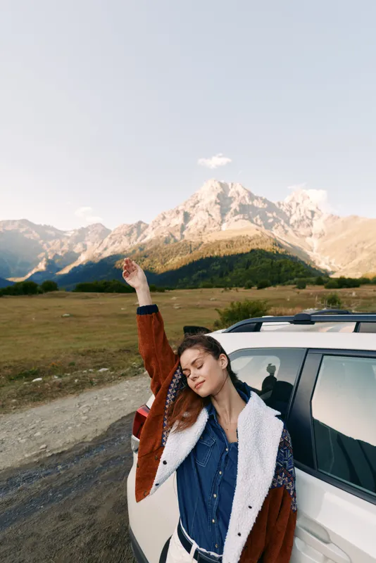 Woman by a car in front of mountains and nature, travel on a gravel road captured as a portrait. Young woman wearing jacket and denim, enjoying sunlight and outdoor freedom during journey.