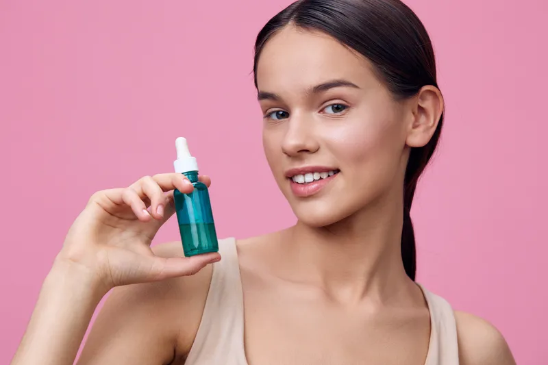 Young woman with flawless skin holding a green serum bottle against a pink background, embodying beauty and skincare concepts in a fresh and vibrant style