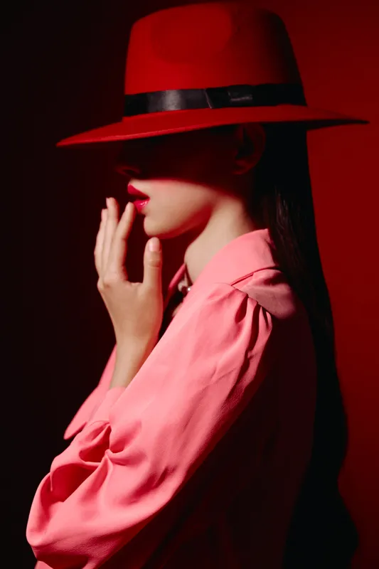 Stylish woman wearing red hat and pink blouse gently touches her lips against red background. Low key lighting highlights dramatic beauty and fashion concept.