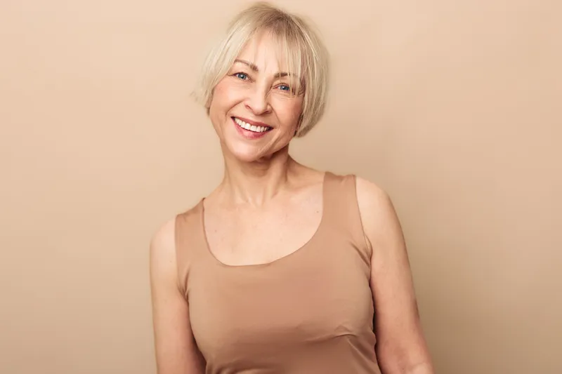 Smiling mature woman with short blonde hair and natural skin poses against beige background wearing sleeveless top. Concept of beauty, confidence, aging gracefully, and positive lifestyle.