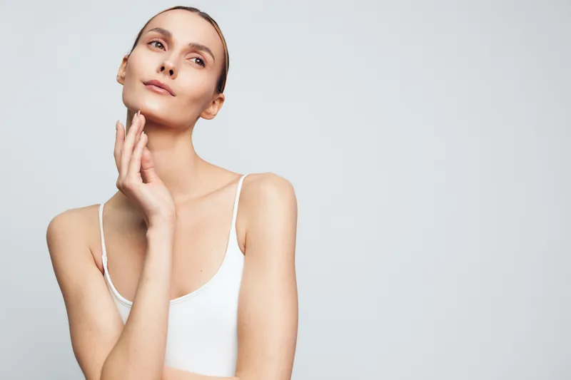 Young woman with clear skin wearing white tank top touching her face with hand. Studio portrait on light gray background. Concept of skincare, beauty, and natural wellness.