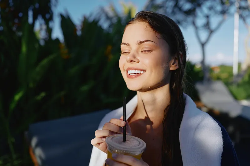 Young woman with closed eyes enjoys sunny morning outdoors holding iced coffee cup with straw wrapped in towel, relaxed lifestyle moment in natural garden setting.
