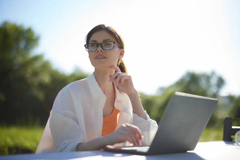 Young woman in eyeglasses working outdoors on a laptop, reflecting a calm and focused mood in a bright, sunny environment surrounded by nature