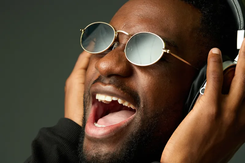 Young Black man wearing sunglasses and headphones, singing passionately against a dark background, evoking joy and musical passion in a modern urban style