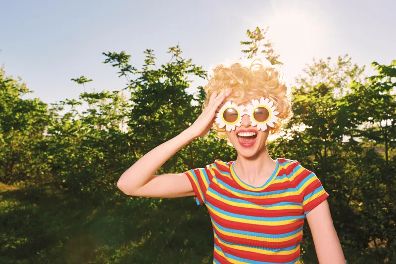 Smiling woman outdoors in a bright striped shirt with flower sunglasses, captured in a retro film color palette. Sunny, playful mood emphasizes carefree lifestyle and summer vitality.